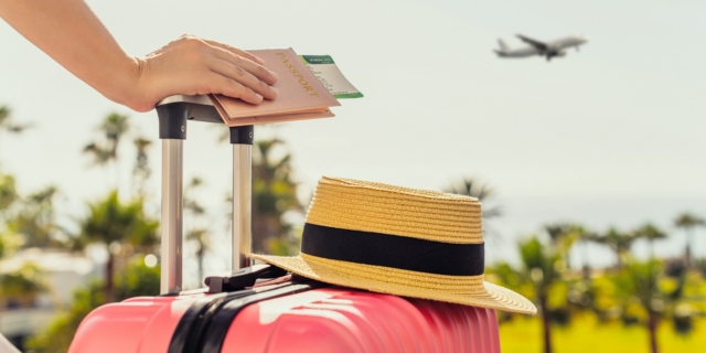 Woman with pink suitcase and passport with boarding pass standing on passengers ladder of airplane opposite sea with palm trees. Tourism concept